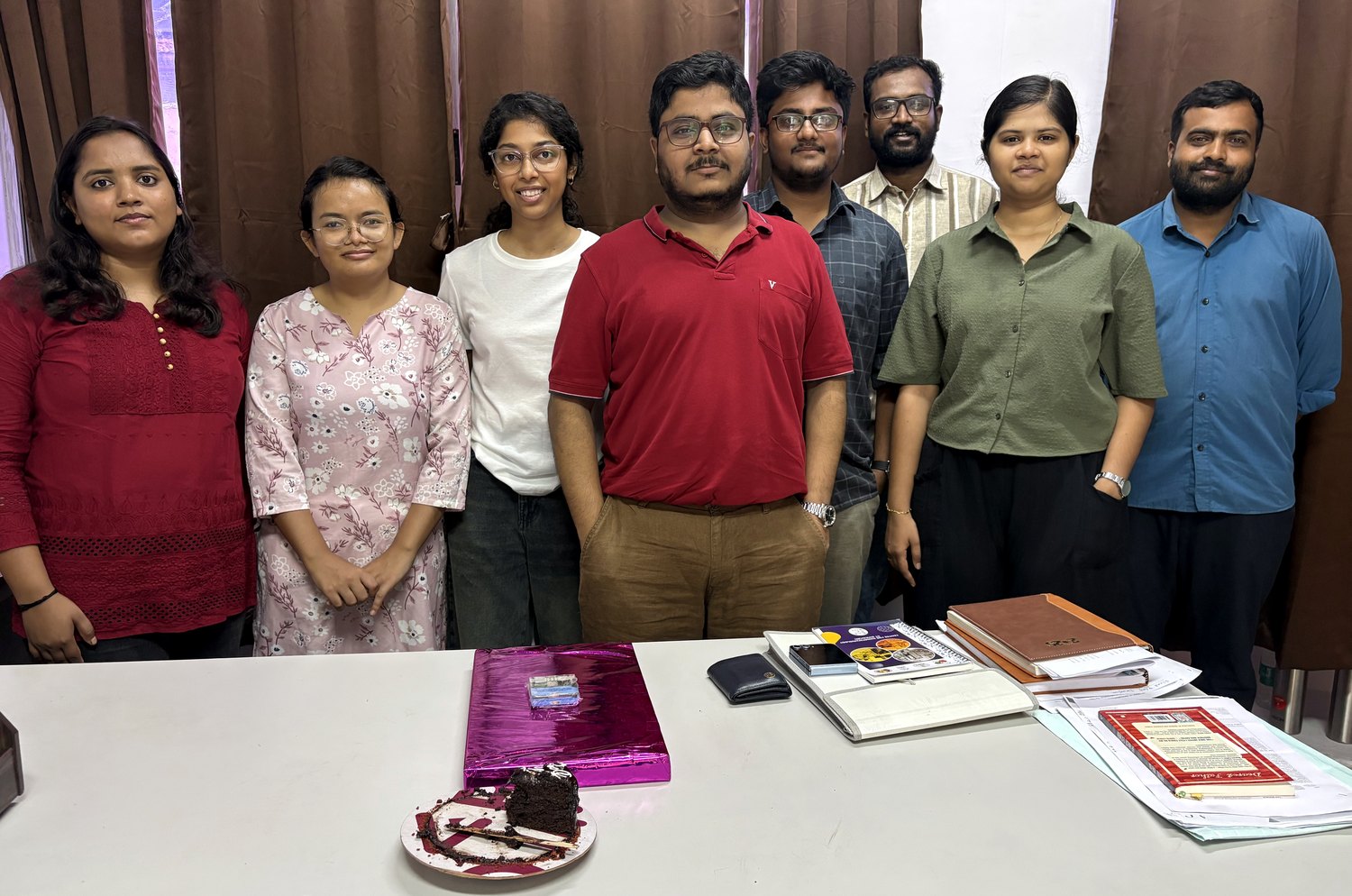 Eight members of the Biosensing and Biophysics Lab standing behind a table with a cake and a wrapped gift, in the PI's office at IIT Guwahati.