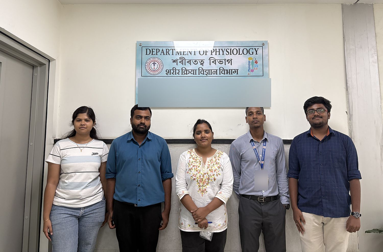 Five members of the Biosensing and Biophysics Lab standing beneath the Department of Physiology signboard at AIIMS Guwahati.