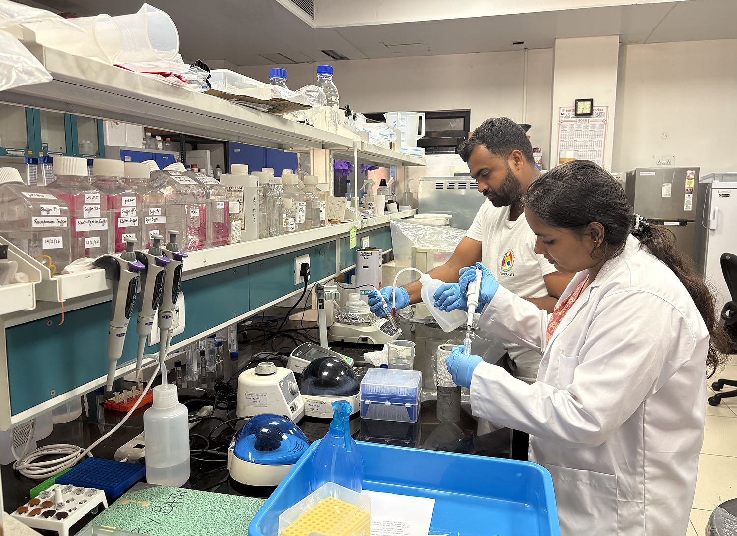 Two researchers in lab coats pipetting at a wet bench lined with buffer stocks, pipettes and microcentrifuges.
