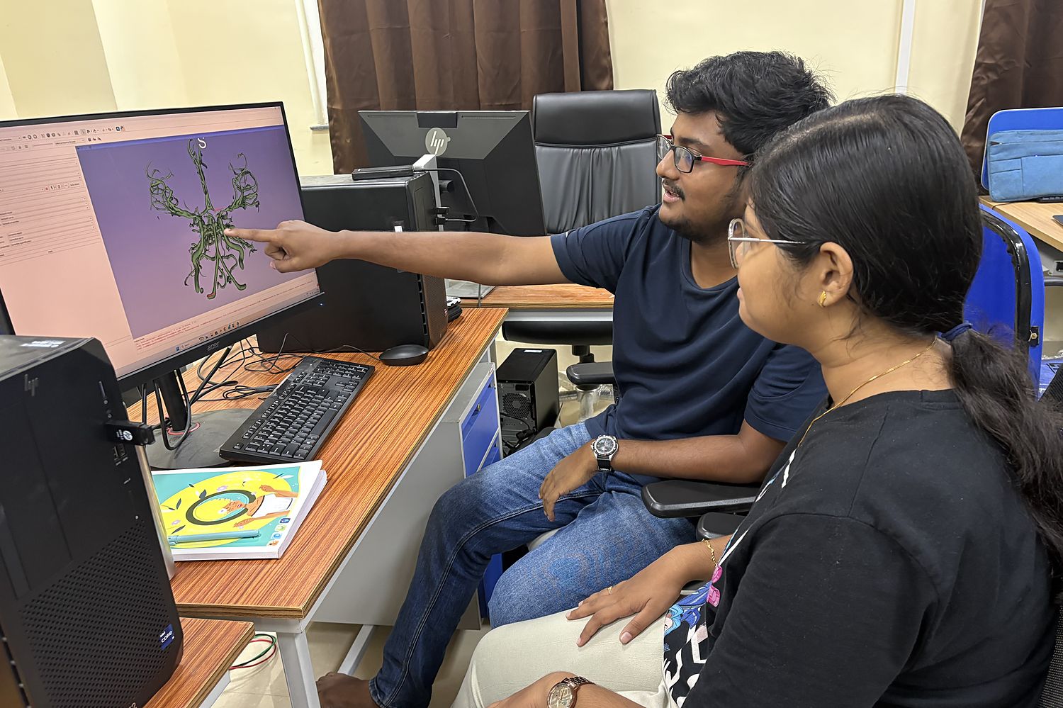 Two members of the lab at a workstation, one pointing at a three-dimensional cerebral arterial tree on the monitor.