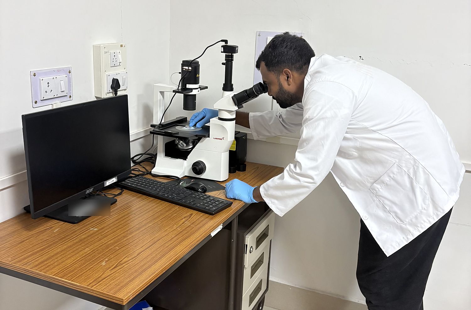 A researcher looking through an inverted microscope fitted with a camera, a sample dish on the stage.