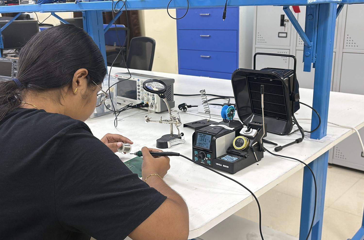 A lab member soldering at a bench fitted with a soldering station, helping-hands stand, oscilloscope and fume extractor.